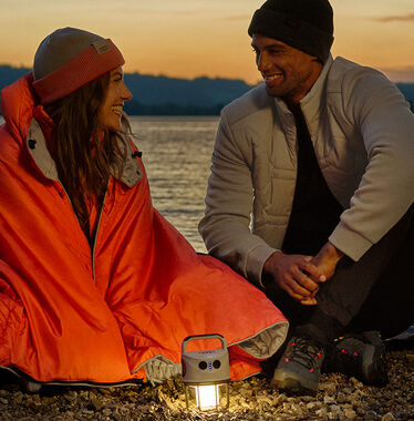Žena s oranžovou dekou, muž v béžové bundě, lampa a termohrnek s logem Audi