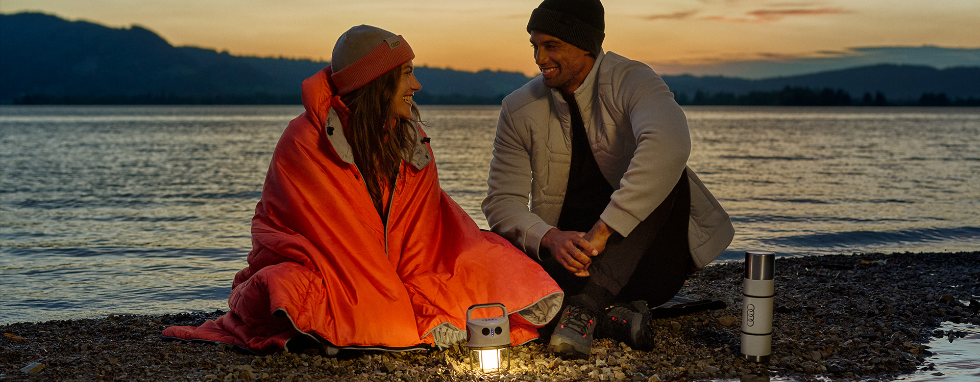 Žena s oranžovou dekou, muž v béžové bundě, lampa a termohrnek s logem Audi