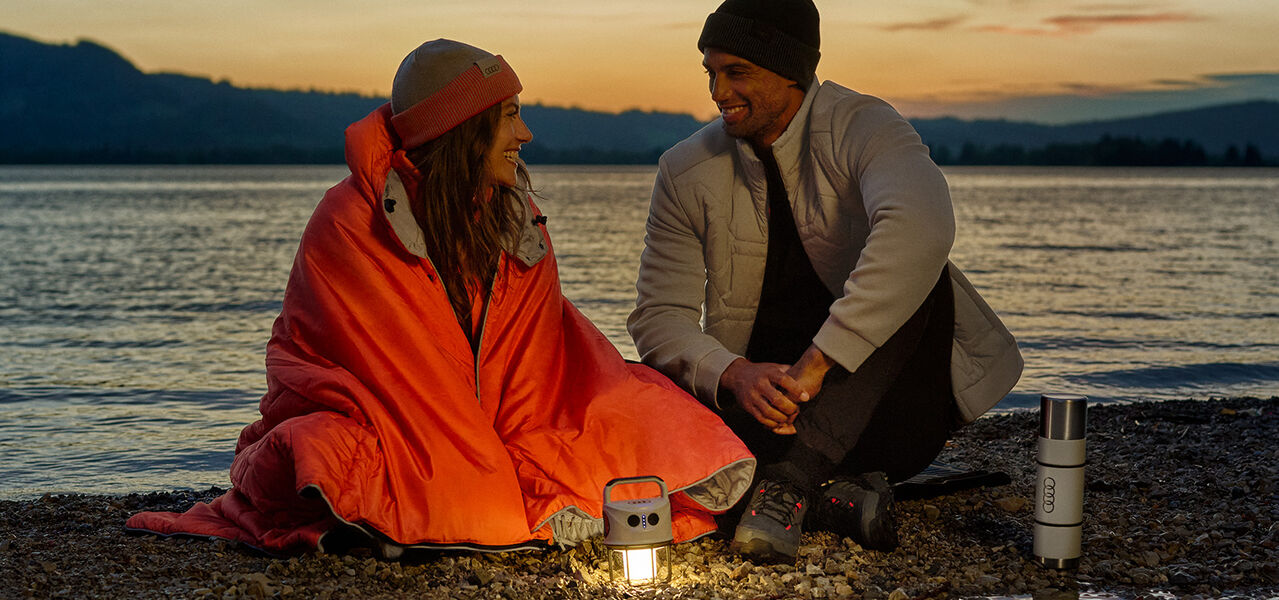 Žena s oranžovou dekou, muž v béžové bundě, lampa a termohrnek s logem Audi