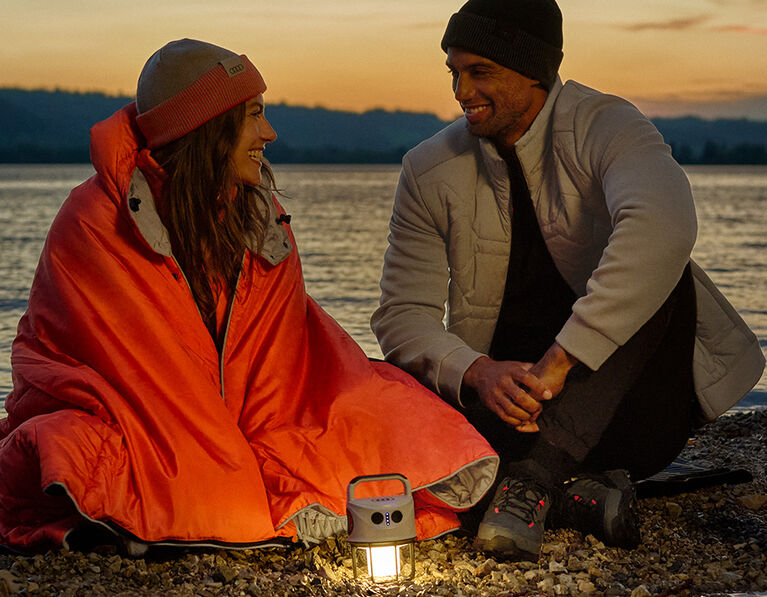 Žena s oranžovou dekou, muž v béžové bundě, lampa a termohrnek s logem Audi